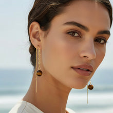 Load image into Gallery viewer, A pair of gold-toned long drop earrings featuring a small chain, a round tiger's eye bead, and a thin dangling wire, shown on a plain white background.