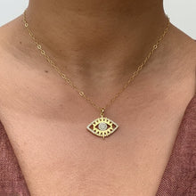 Load image into Gallery viewer, A gold chain necklace with an evil eye pendant worn by a person, shown from the chest up against a brick wall background.
