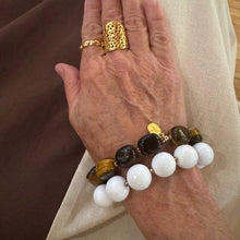 Load image into Gallery viewer, A close-up of a person's hand resting on a beige fabric surface, wearing a beaded bracelet with white, dark brown, and tiger's eye beads, along with gold rings.
